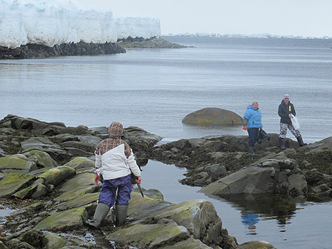 Three people are standing on the rocky shore by the water. All of them are dressed with rubber boots and winter clothes. One of them is holding a long knife and the other two are holding plastic bags. In the background, we see the expanse of water bordered by rocky shores on which rests a high ice floe.