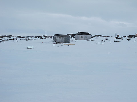 Winter landscape, cloudy sky: two small huts are set up on top of a rocky hill where only the peaks are covered with snow. They seem to be made of used wooden panels.