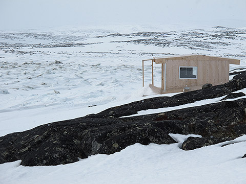 In a hilly and rocky landscape, mainly covered with snow and ice, a wooden cabin appears to have been recently installed. It’s composed of natural coloured wooden panels not yet greyed by the sun.
