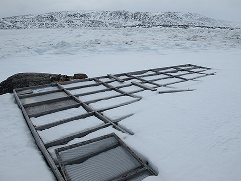 On the shores of a frozen creek bordered by a partially snow-covered rocky mountain, a wooden platform begins to appear due to the melting of the snow that covered it. This platform is divided into rectangular boxes. A wire frame rests on the platform in the foreground.