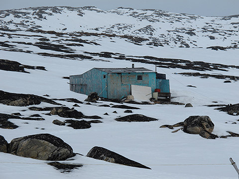 A cabin, in a partially snow-covered rocky hill. It’s made of used wooden panels with faded blue paint. Some objects and building materials are placed around the cabin.