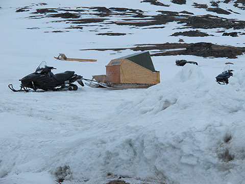 A covered sled is attached to a snowmobile. It’s made of pieces of wooden panels and sheet metal, and represents a small enclosed cab on skis. There’s snow on the ground all around. Another snowmobile and a toboggan rest nearby. Two off-road vehicles are revealed by the melting snow. They're still half-buried. In the background, we can see a rocky hill partially covered with snow.